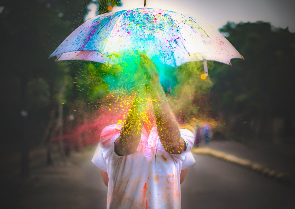 An image of a person in a white shirt standing under an umbrella. They are clapping their hands and rainbow colors are flying all over.